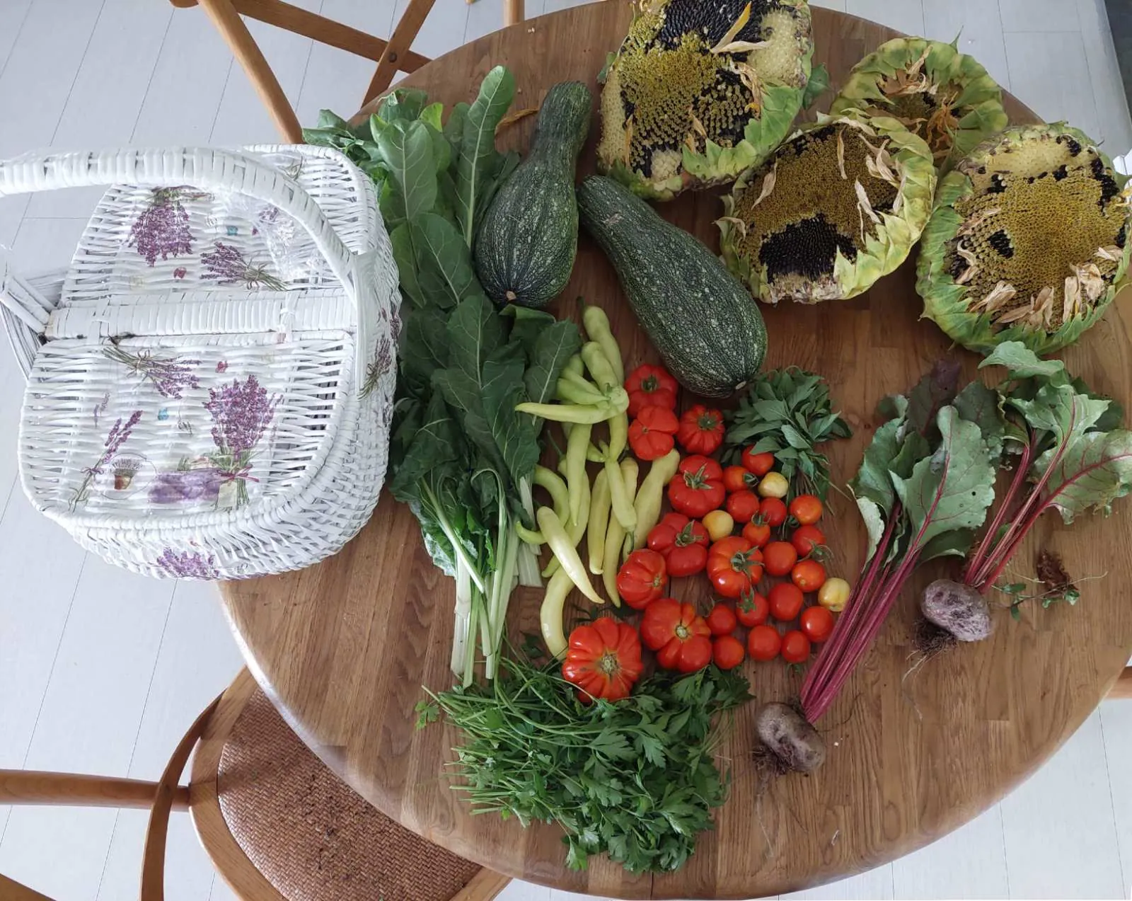 A late-summer garden harvest arranged on a round wooden table — courgettes, ripe tomatoes, pale peppers, beetroot with its leaves, herbs and cut sunflower heads beside a white wicker basket