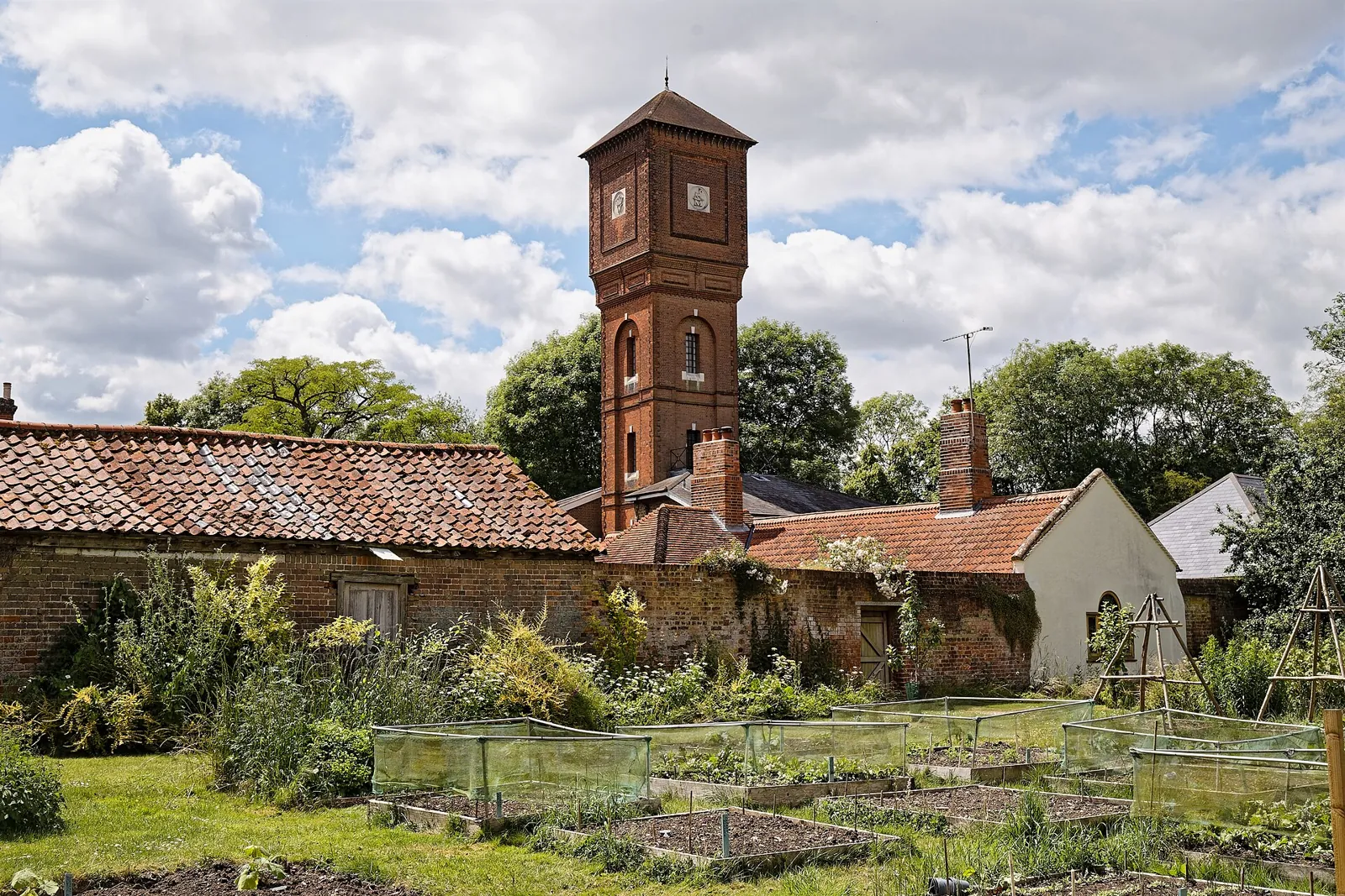 Square vegetable beds under low crop-protection cages in a walled kitchen garden, a Victorian brick water tower rising behind the wall